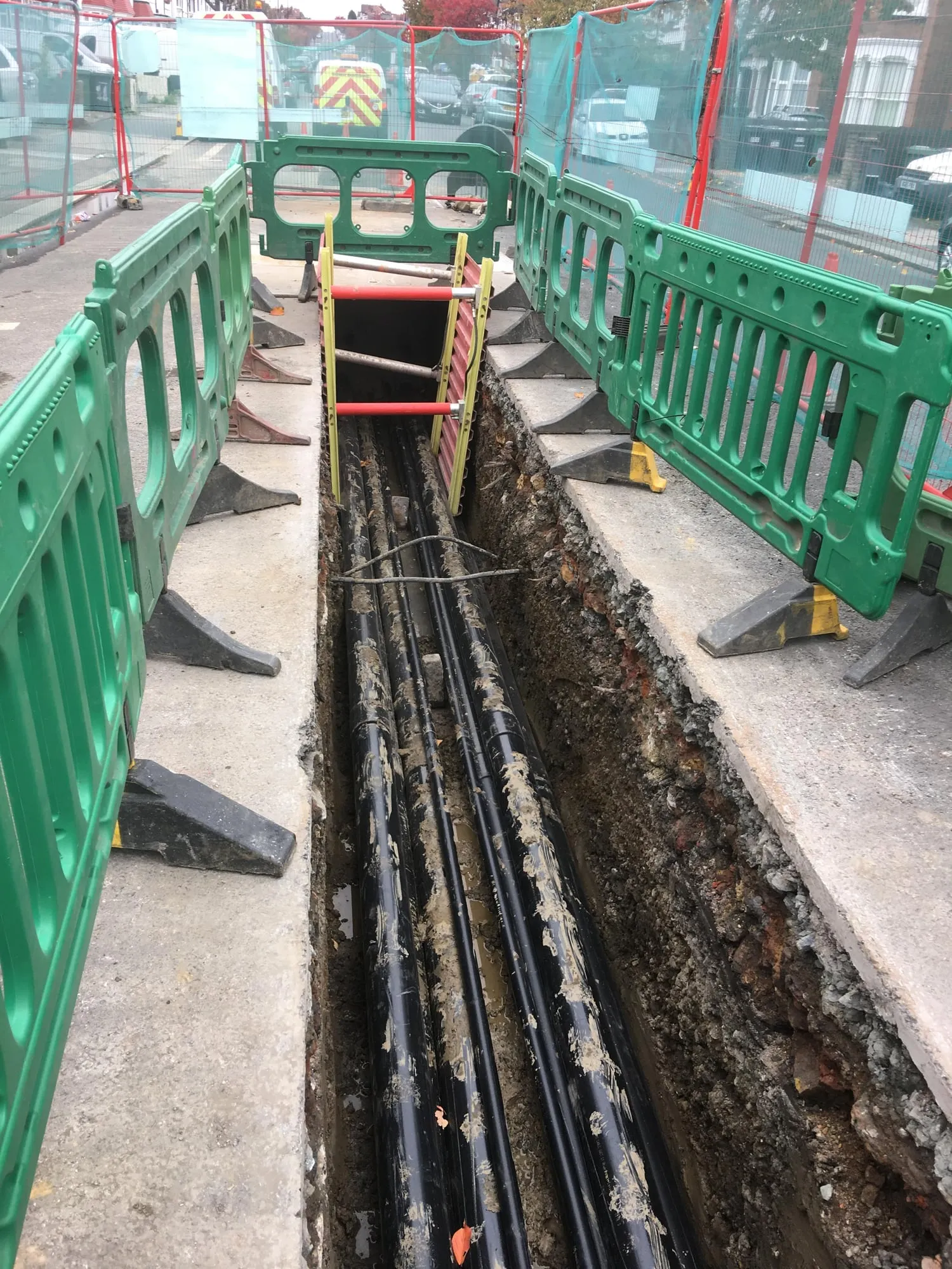 Open utility trench with new black ducts laid, behind green barriers